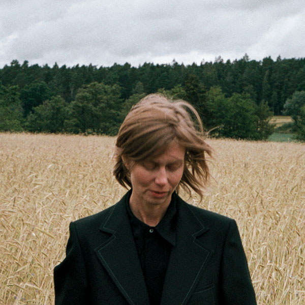 Eine Person mit kurzem Haar steht in einem Getreidefeld und blickt nach unten. Wind bewegt das Haar, im Hintergrund ein Wald unter bewölktem Himmel.