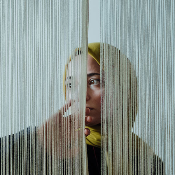 Porträt einer Person mit gelbem Kopftuch, die hinter dünnen Fäden oder Vorhängen hervorschaut und eine Hand davor hält.