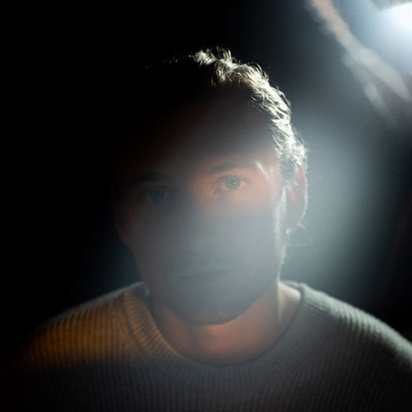 Dunkel belichtetes Portrait eines Mannes mit leichtem Bart, der in Richtung Kamera blickt. Ein Lichtstrahl fällt quer über sein Gesicht.