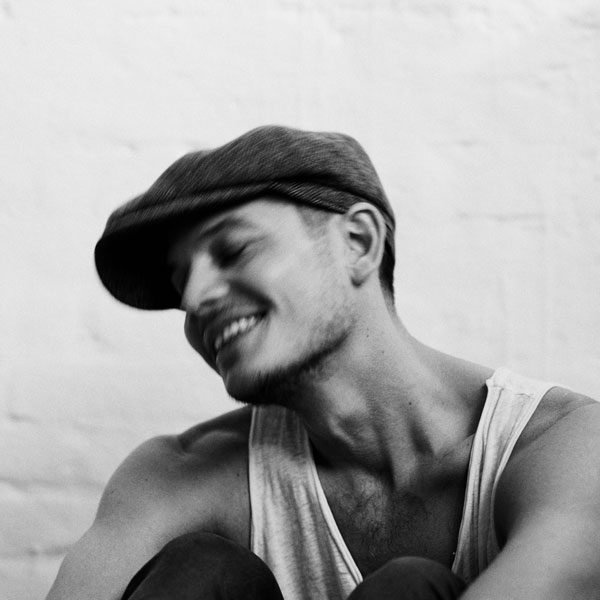 Schwarzweißfoto eines lächelnden Mannes mit Schiebermütze und Trägershirt, der leicht zur Seite geneigt sitzt.