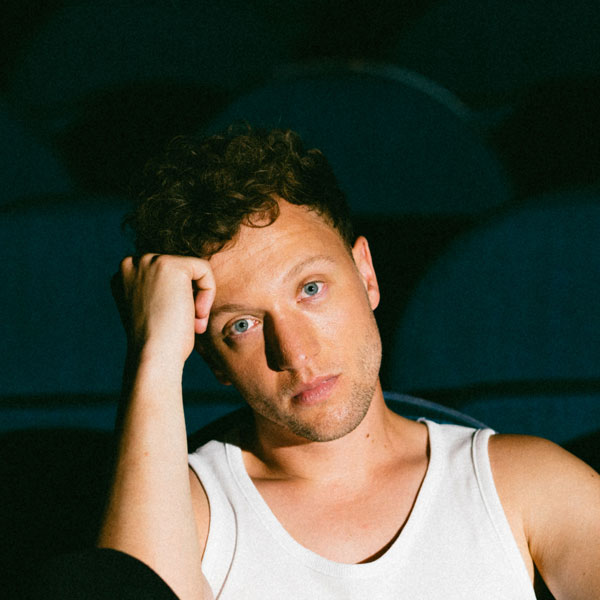 Portrait eines Mannes in einem weißen Tanktop, der den Kopf in die Hand stützt und nachdenklich in die Kamera schaut.