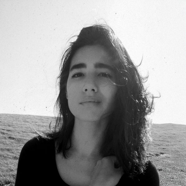 Schwarzweißfoto einer Frau mit mittellangem Haar, die ruhig in die Kamera schaut. Im Hintergrund eine offene Landschaft und heller Himmel.