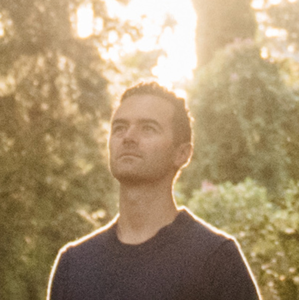 Eine Person steht im Gegenlicht draußen, umgeben von Bäumen und warmem Sonnenlicht. Blick leicht nach oben gerichtet.