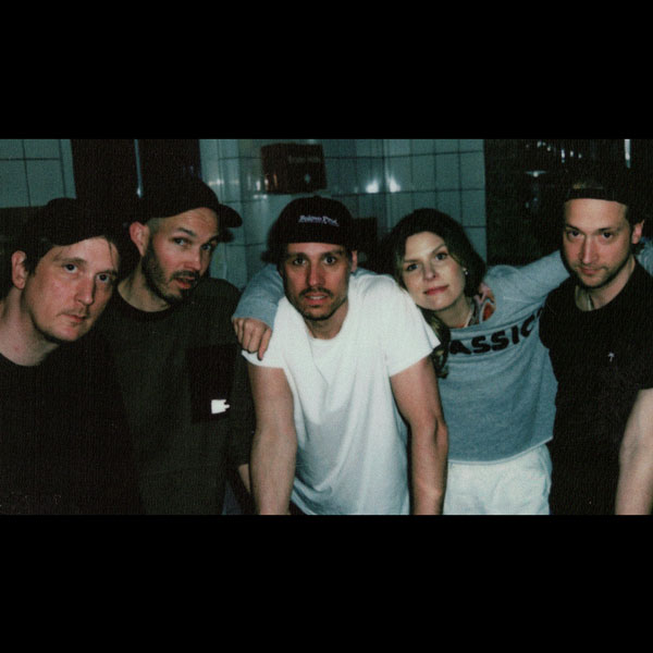 Fünf Personen stehen eng zusammen in einem Innenraum. Sie lehnen sich freundschaftlich aneinander und schauen in die Kamera.