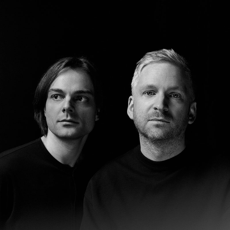 Schwarzweißportrait von zwei Personen vor dunklem Hintergrund. Beide tragen dunkle Kleidung; eine Person blickt leicht zur Seite, die andere direkt in die Kamera.