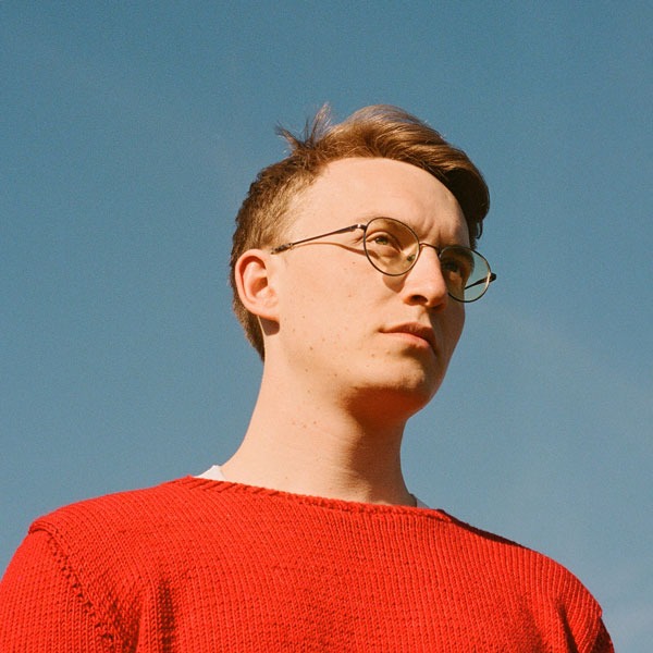 Person mit kurzem, hellbraunem Haar und runder Brille, trägt einen roten Strickpullover und blickt ernst in die Ferne. Klarer blauer Himmel im Hintergrund.