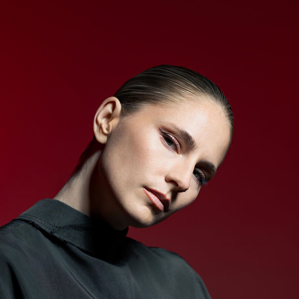 Porträt einer Person vor rotem Hintergrund, mit streng nach hinten gekämmtem Haar und leicht geneigtem Kopf. Ruhiger, intensiver Gesichtsausdruck.