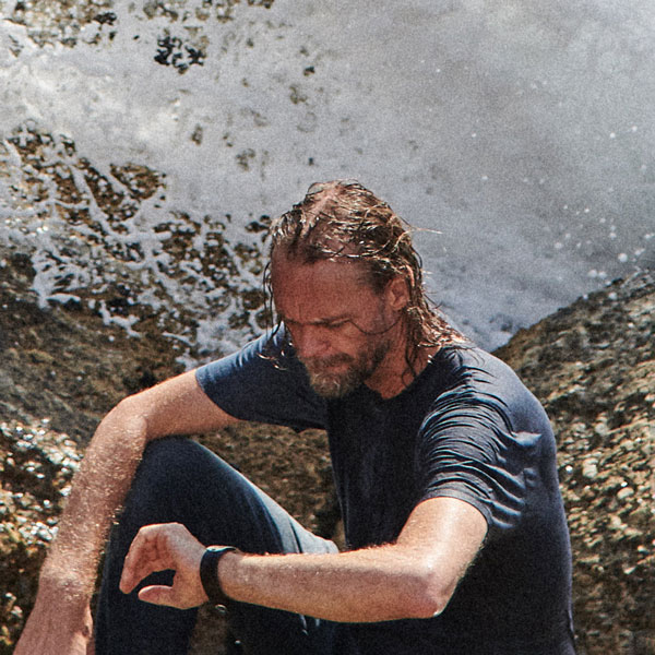 Person mit nassem Haar und nassem T-Shirt sitzt auf einem Felsen am Meer, während eine Welle im Hintergrund spritzt. Der Blick ist nach unten gerichtet.