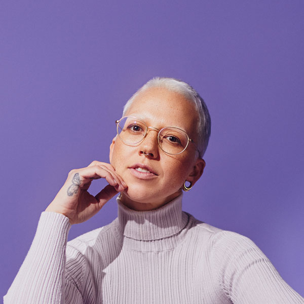 Porträt einer Person mit rasiertem, hellblondem Haar, runder Brille und hellem Rollkragenpullover vor lilafarbenem Hintergrund. Eine Hand liegt locker am Kinn.“