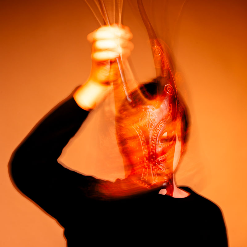 Stark verschwommenes, orangefarbenes Bild einer Person, die eine verzierte Maske hält oder trägt. Die Bewegung erzeugt einen dynamischen, abstrakten Effekt