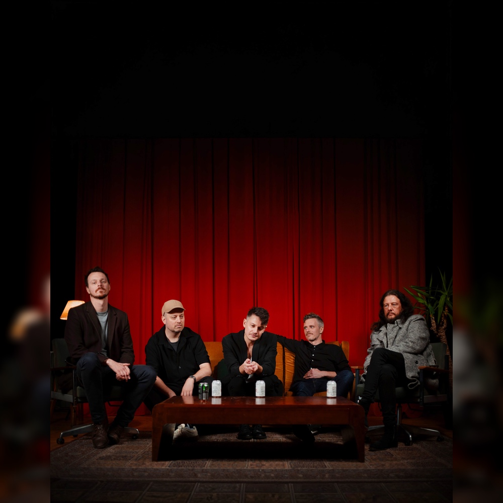 Farbliches Bild in rot schwarz mit fünf Männern, die auf einem Sofa und auf Stühlen vor einem Tisch sitzen